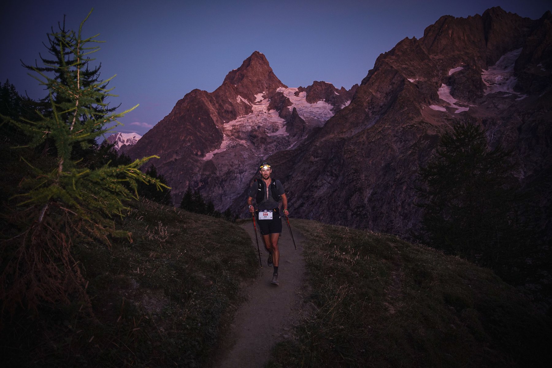 Jack Chamberlain climbing a trail by headtorch at first light during UTMB Mont-Blanc, with the Aiguille Noire lit pink behind him.